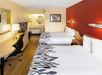 A hotel room with two double beds, a TV, a desk, a chair, and a lamp. The walls are red and cream, with a framed picture above. The floor is wooden.