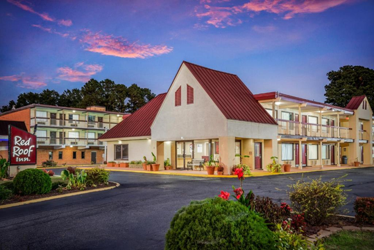 A Red Roof Inn motel with a red-roofed building and a two-story structure, surrounded by plants and a parking area, under a sky with pink clouds.