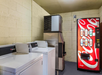 A laundry room featuring two washing machines, an ice machine, and a Coca-Cola vending machine against a beige brick wall.