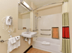 Hotel bathroom with a wall-mounted hairdryer, sink, mirror, bathtub with grab bars, handheld showerhead, and a wooden seat. Towels are hung near the sink and bathtub.