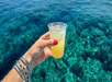 A hand with red nail polish and silver bracelets holds a plastic cup with a yellow drink over clear, blue ocean water.