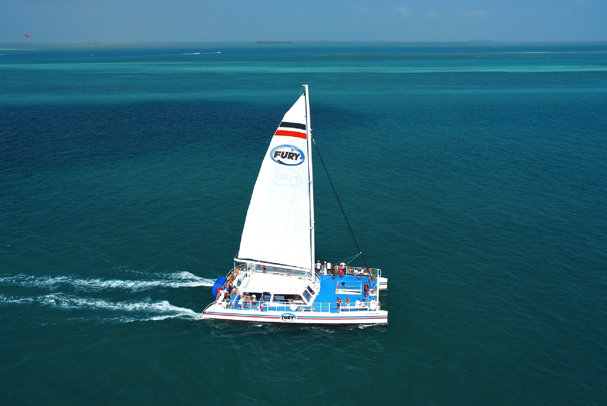 A catamaran with a white sail labeled "Fury" sails on a calm, blue ocean with a group of people onboard.