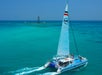 A catamaran sails through clear turquoise waters, with a distant buoy visible on the horizon under a clear blue sky.