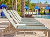 A row of empty lounge chairs is lined up beside a swimming pool, with blue cabanas and palm trees visible in the background on a sunny day.