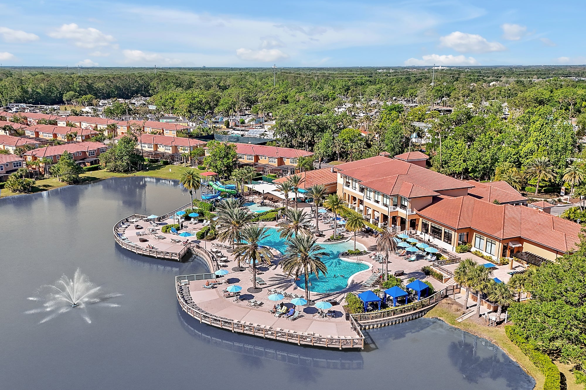 Aerial view of a lakeside resort with a pool, sun loungers, palm trees, a water fountain, and surrounding residential buildings amid greenery.