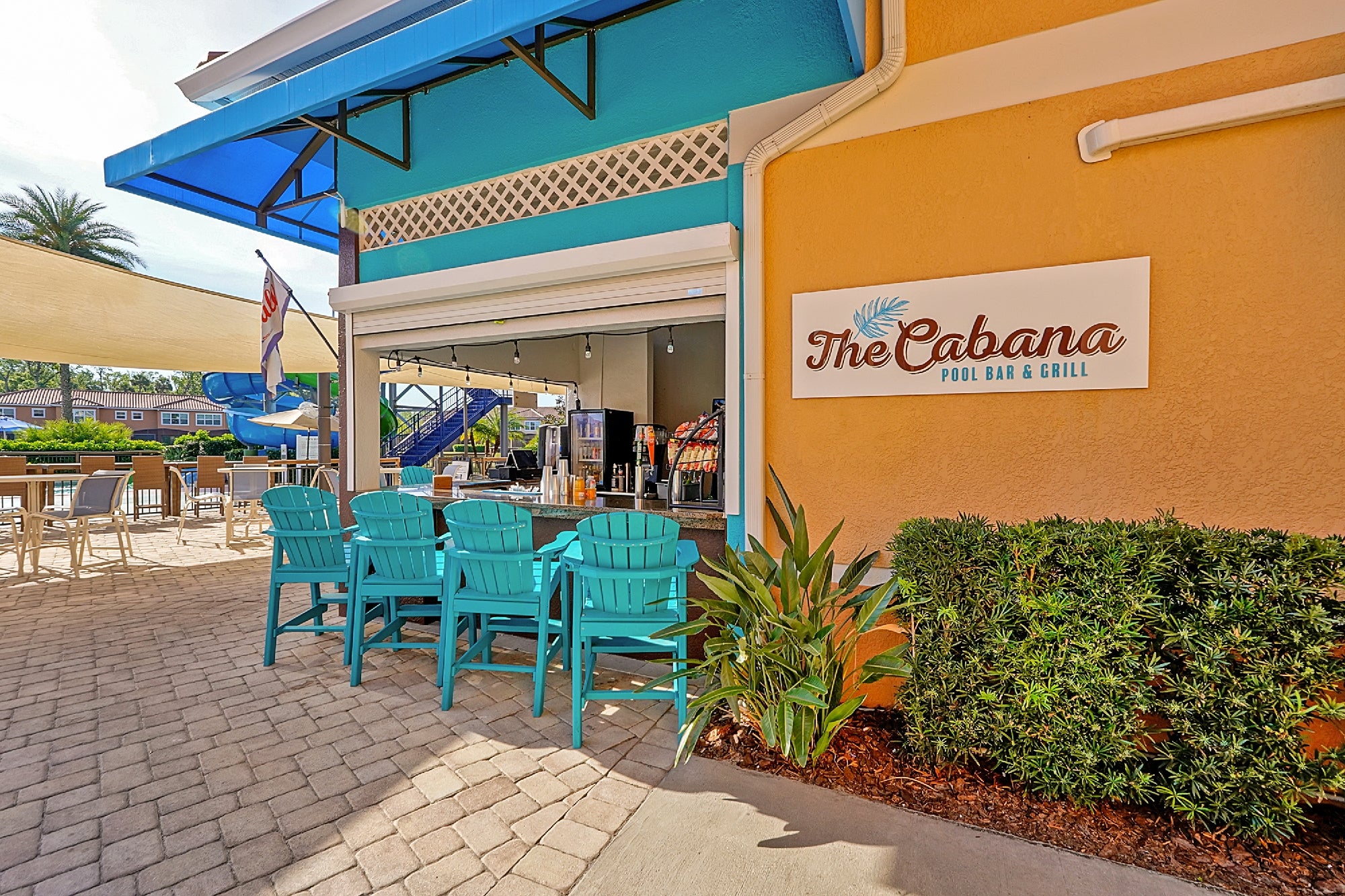 Outdoor poolside bar called "The Cabana" with turquoise chairs lined up at the counter, tropical plants, and a shaded seating area.