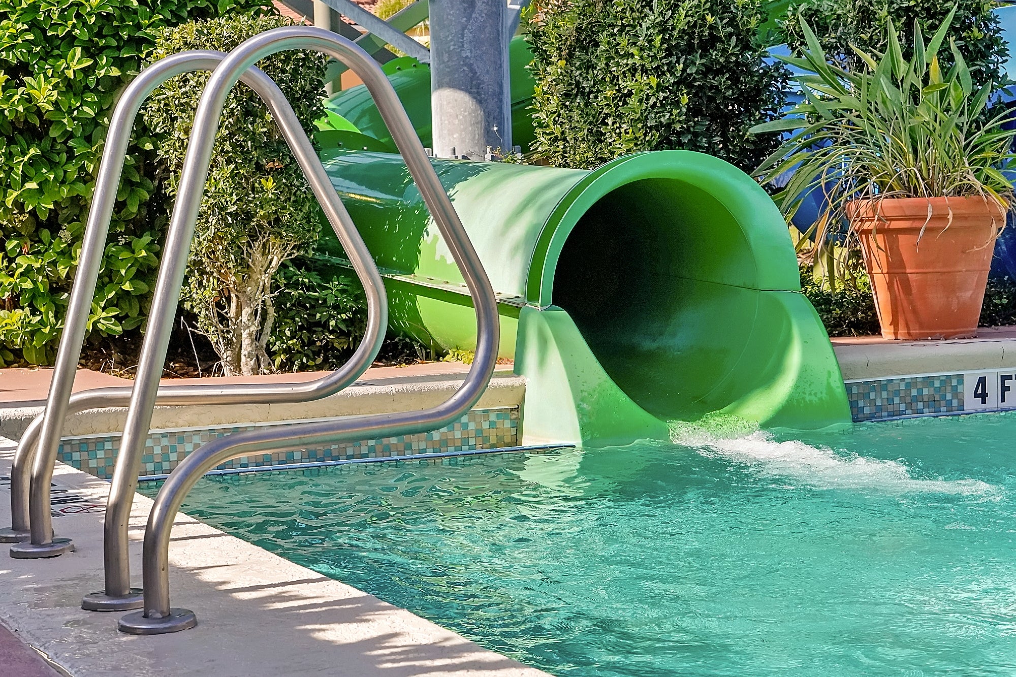 A green water slide ends in a swimming pool with clear water; metal handrails and potted plants are visible nearby.