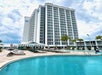 A tall hotel building stands behind a pool with lounge chairs, umbrellas, and palm trees under a cloudy sky.