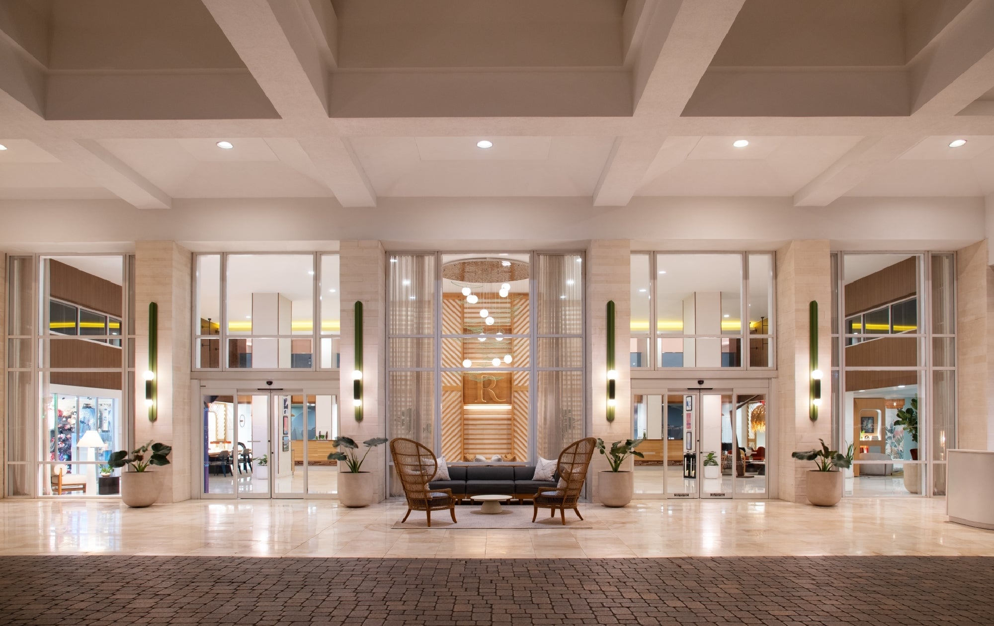 Covered entrance of a modern building with glass doors, potted plants, a small seating area with two chairs and a sofa, and warm overhead lighting.