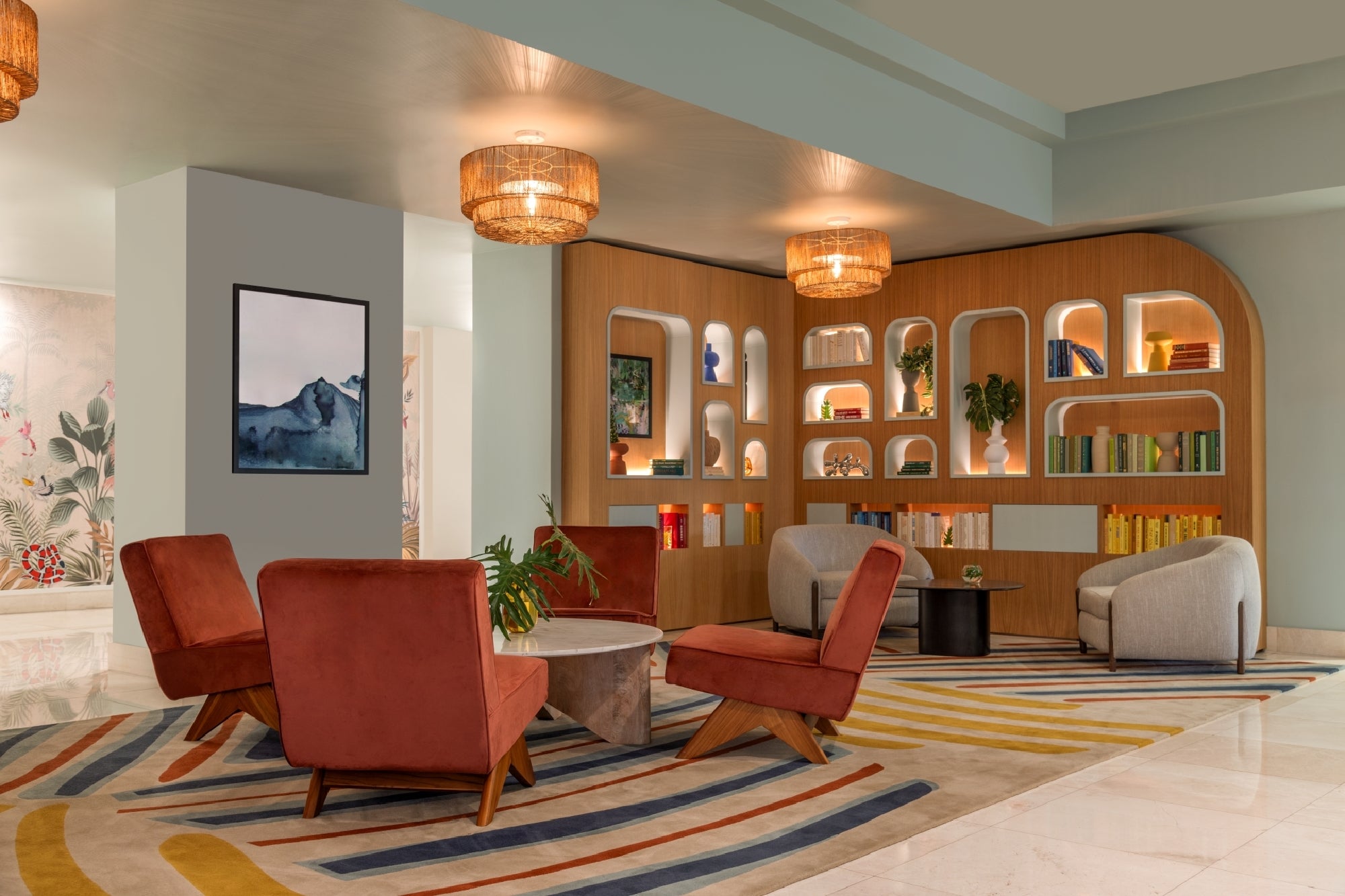 Modern lounge area with red and gray chairs, a round table, bookshelves with decor, art on the wall, and a patterned rug on a tiled floor. Warm lighting from ceiling fixtures.