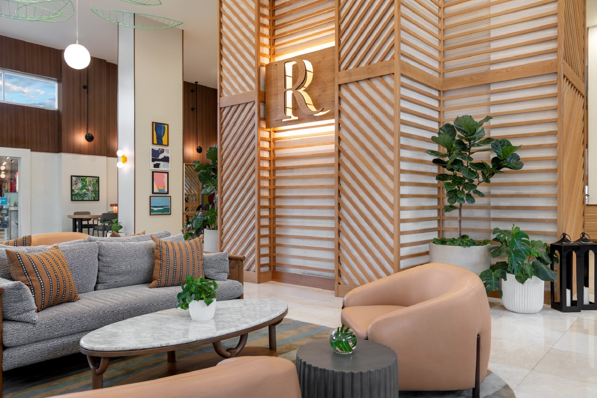 Modern hotel lobby with gray sofa, tan chairs, marble coffee table, potted plants, and a large wooden accent wall featuring a prominent letter "R" sign.