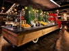 A restaurant buffet station styled as a vintage red truck, offering pastries, fruit, and cereals, with greenery and exposed lighting in the background.