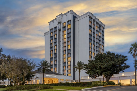 A tall, modern hotel building with large windows, "Renaissance Orlando" signage, palm trees, and a partly cloudy sky at sunrise or sunset.
