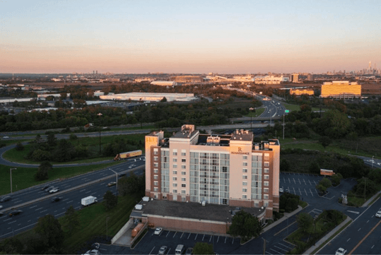 Aerial view of a Residence Inn by Marriott hotel surrounded by roads, green areas, and industrial buildings with a distant city skyline under a clear sky.