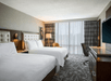 Hotel room with two large beds, a flat-screen TV, desk, and chair. Floor-to-ceiling window with drawn sheer curtains. Neutral decor and patterned carpet.