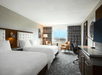 A hotel room with two double beds, a TV, desk, and chairs. Large window with view outside. Neutral tones and contemporary decor.