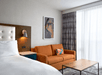 Hotel room with a white bed, orange sofa, wooden coffee table, abstract wall art, and large window with curtains.