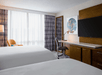 Hotel room with two beds, a desk with a chair, large TV, floor lamp, and artwork. A window with sheer curtains allows natural light to enter the room.