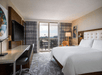 Hotel room with a king-size bed, desk, TV, and balcony view. Neutral decor with modern furnishings and a large window allowing natural light.