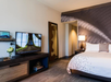 Stylish hotel room with a large bed, wall-mounted TV, modern decor, and a standing lamp. The room includes a patterned wall and a tall mirror beside the bed.