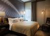 A hotel room with a large bed, decorative pillows, a geometric wall pattern, bedside lamps, a chair, and sheer curtains over a window.