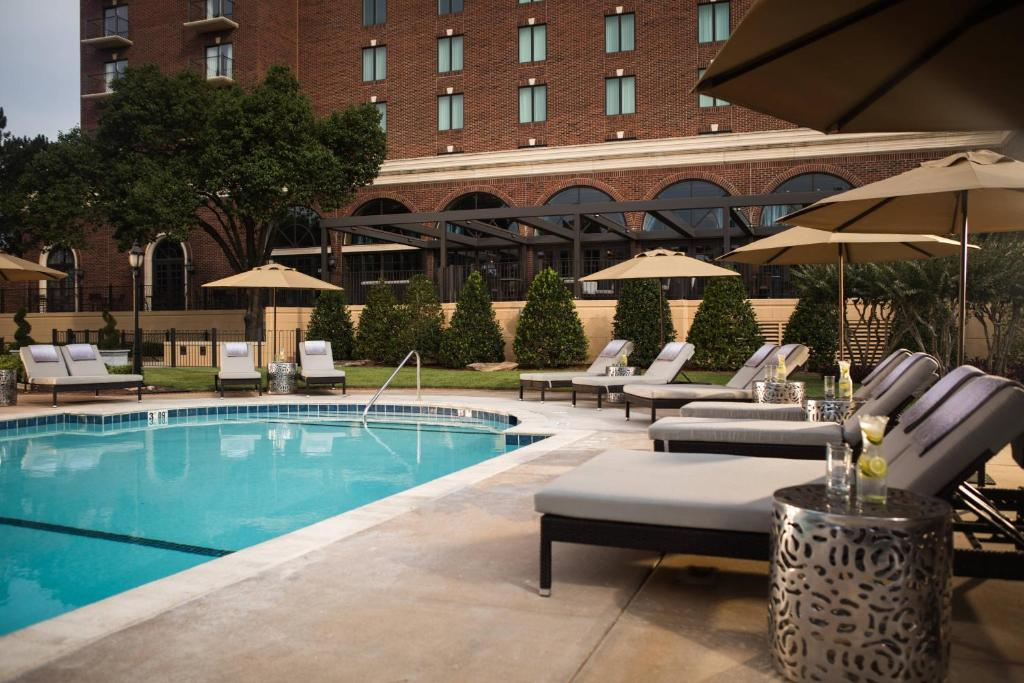 Outdoor hotel pool area with lounge chairs, umbrellas, and a brick building in the background. Tables with drinks are next to the chairs.