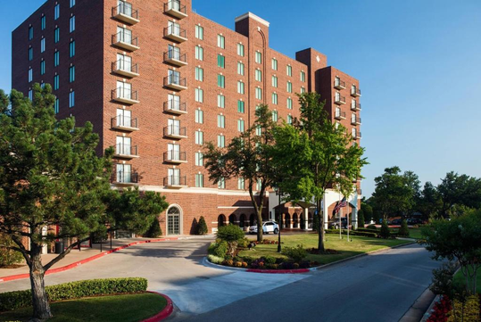 Red-brick hotel with balconies, surrounded by trees and a driveway. American flag visible at entrance. Brightly lit on a clear day.