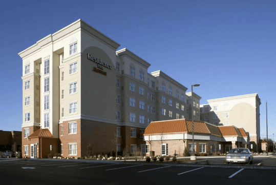 A multi-story hotel building with a beige facade and white accents under a clear blue sky, featuring numerous windows and a small entrance area with a brown roof.