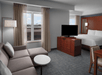 Contemporary hotel room with a bed, sofa, and flat-screen TV. Large window with sheer curtains allows natural light, revealing a city view. Neutral tones and modern furnishings complement the space.