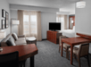 A hotel room with a seating area, flat-screen TV, bed, curtains, and wooden furniture, including a desk and dining table with chairs.