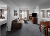 Hotel suite featuring a living area with a sofa, armchair, fireplace, and TV, a dining table with chairs, and two bedrooms with beds, all in a neutral color palette.