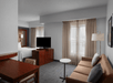 A hotel room with a bed, a TV on a wooden stand, a brown sofa, a round table, and a floor lamp. The space features light grey walls and two windows with beige curtains.