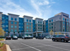 A modern five-story hotel with multiple windows and a blue-gray facade is shown. Several cars are parked in the spacious lot in front.