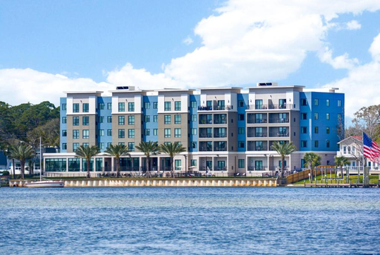 A modern four-story apartment building by a body of water, with balconies, palm trees, and a flagpole.