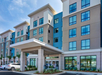 Modern hotel exterior with four stories, large entrance canopy, and multiple windows. Parking lot and landscaping in the foreground.