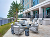 Outdoor seating area with cushioned chairs around a fire pit, near grills and lounge chairs, adjacent to a modern building overlooking a waterfront with palm trees.