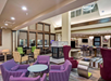 Hotel lobby with modern décor, purple and beige seating, a central fireplace, and a high ceiling with large windows.