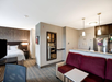 A modern hotel room featuring a bed, sofa, and kitchenette with a fridge. The room is well-lit with pendant lights and neutral decor.
