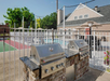 Two stainless steel grills on a stone counter near a fenced area with a pool, surrounded by trees and a building in the background, under a clear sky.