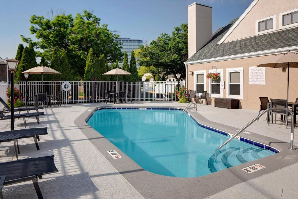 Outdoor swimming pool with a concrete deck, surrounded by lounge chairs and umbrellas. A building with a sloped roof is in the background, and trees are visible beyond the pool fence.