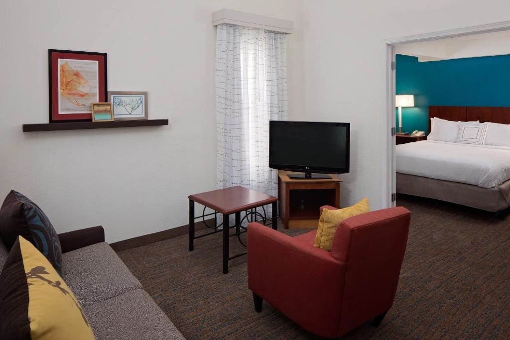 Hotel room with a sitting area featuring a sofa, red chair, TV, and side table. A doorway leads to a bedroom with a bed. Walls are white; one wall in the bedroom is teal.