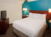 Hotel room with a queen bed, white bedding, and teal accent wall. Two lamps are beside the bed on wooden nightstands. A flat-screen TV is on a dresser to the left.