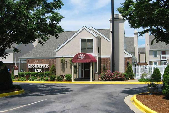 Exterior of a Residence Inn with a red awning, surrounded by landscaped gardens, trees, and parked cars. A "Residence Inn" sign is visible on the left.