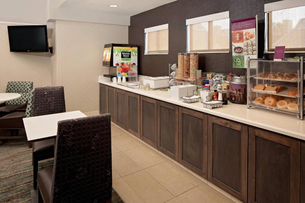 Hotel breakfast area with a counter offering cereal dispensers, a juice machine, pastries, and condiments. Tables and chairs are set for guests.