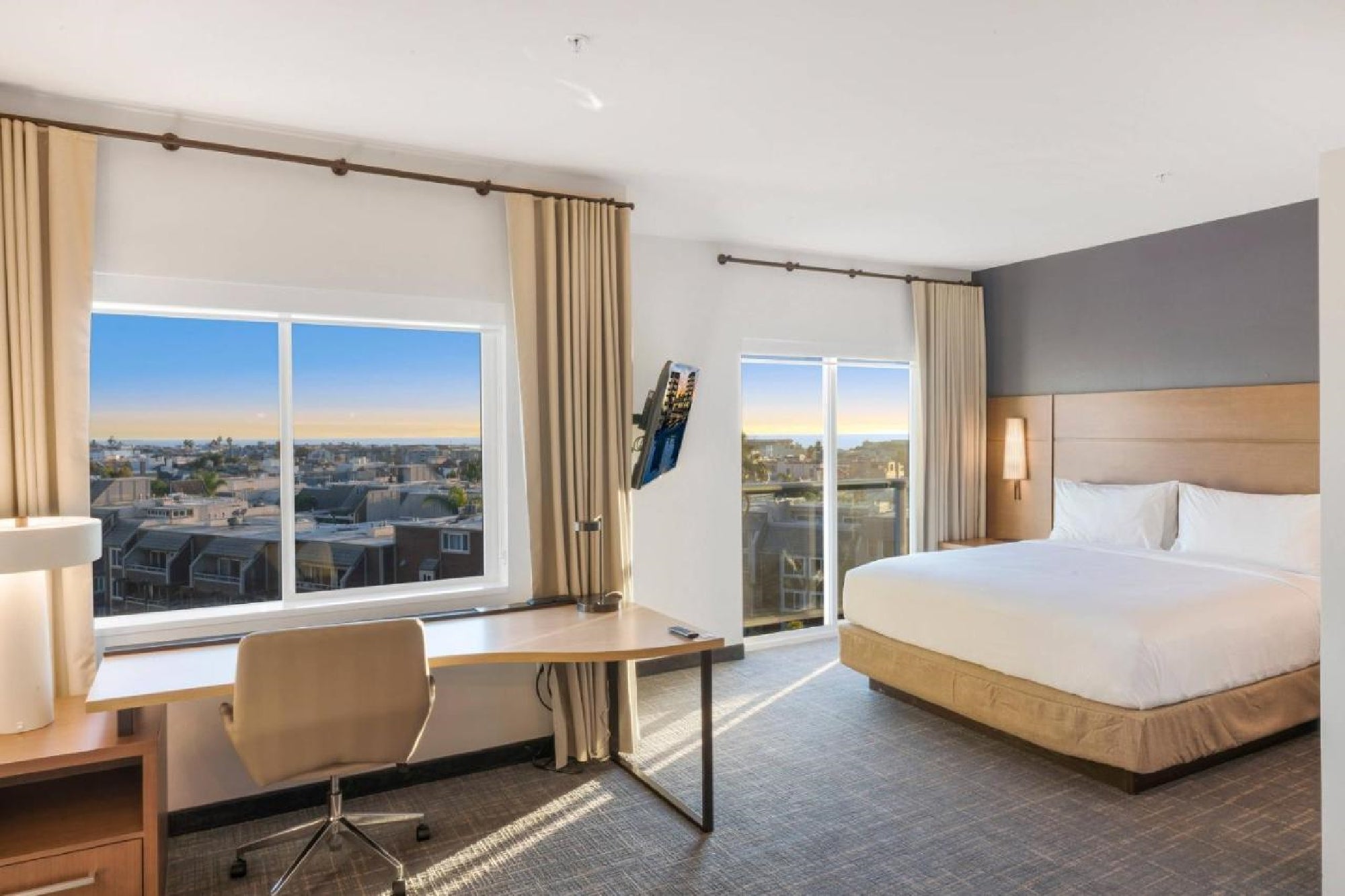 Hotel room with a large bed, desk, chair, and two windows showing a cityscape and sunset. Modern decor with neutral tones.