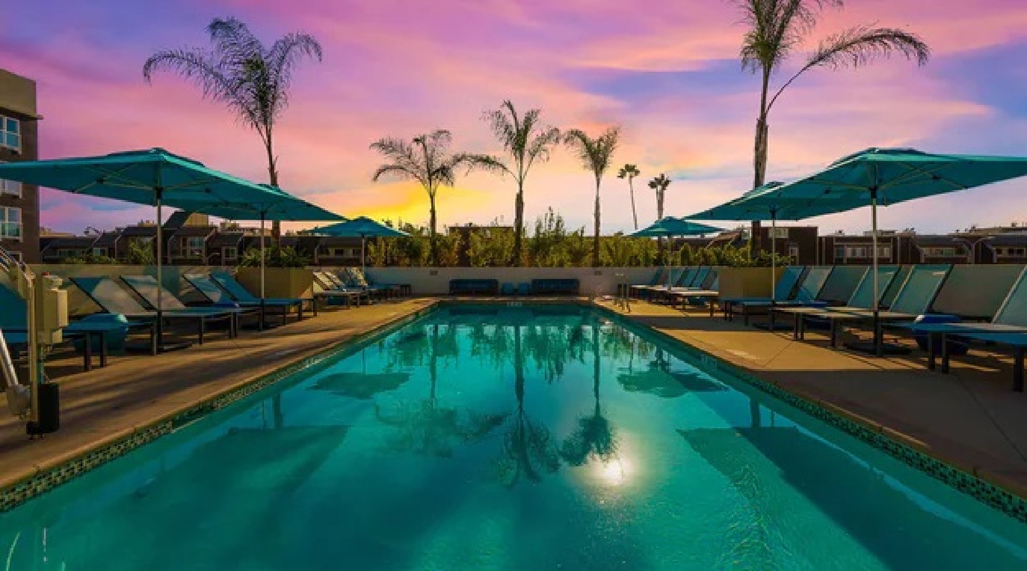 A serene swimming pool surrounded by lounge chairs and umbrellas at sunset, with palm trees silhouetted against a vibrant sky.