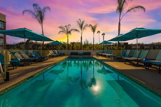 A serene swimming pool surrounded by lounge chairs and umbrellas at sunset, with palm trees silhouetted against a vibrant sky.