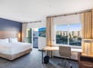 Hotel room with a large bed, desk with chair, lamp, and a window view of nearby buildings. A wall-mounted TV and framed artwork are also visible.