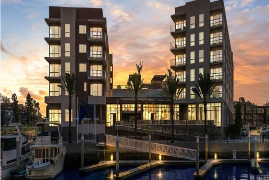 Two modern apartment buildings by a marina at sunset, viewed from across the water, with boats docked and palm trees lining the entrance.
