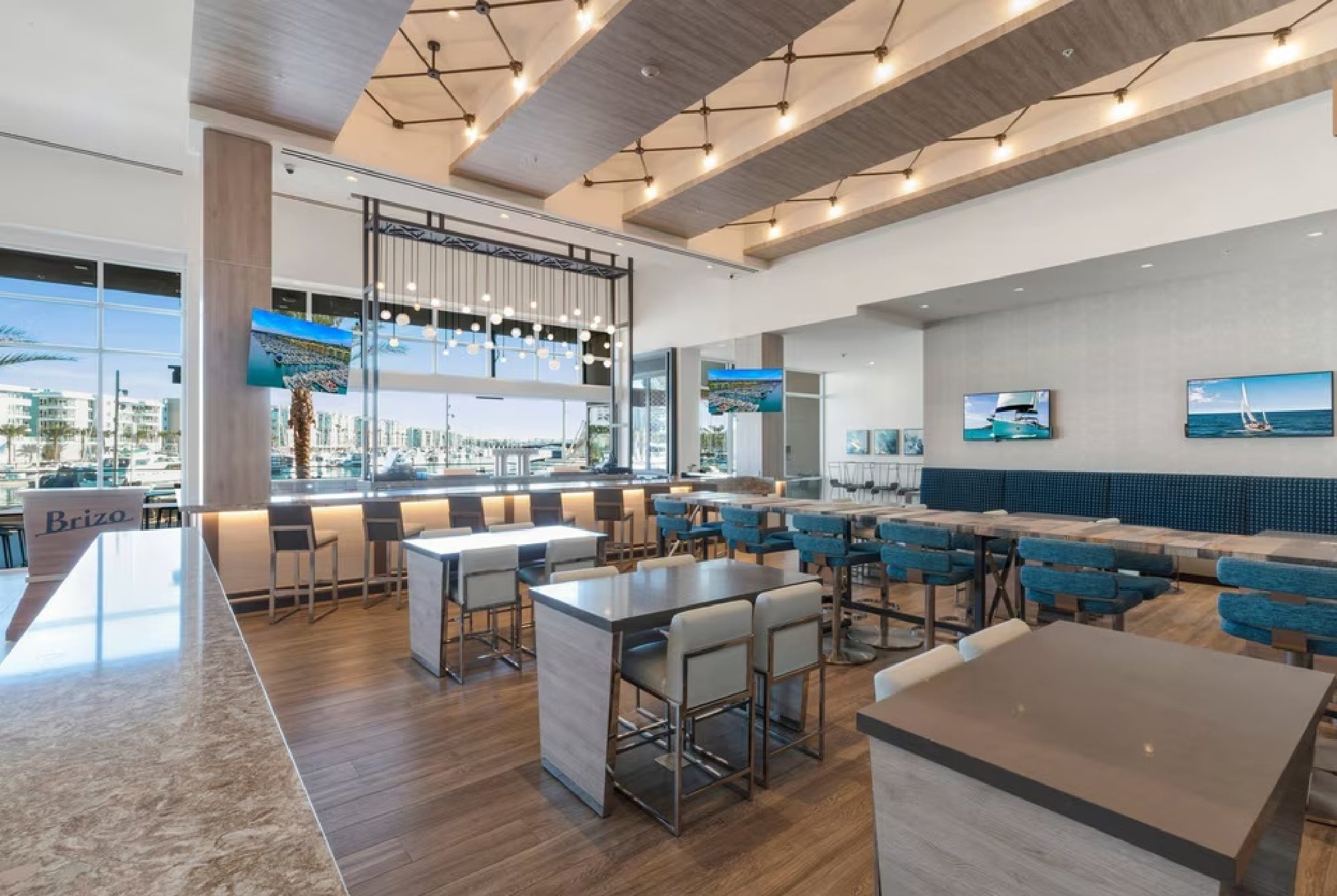 Modern restaurant interior with wooden tables, teal and white chairs, a marble counter, and large windows. Multiple TV screens are mounted on the walls, showing outdoor scenes.